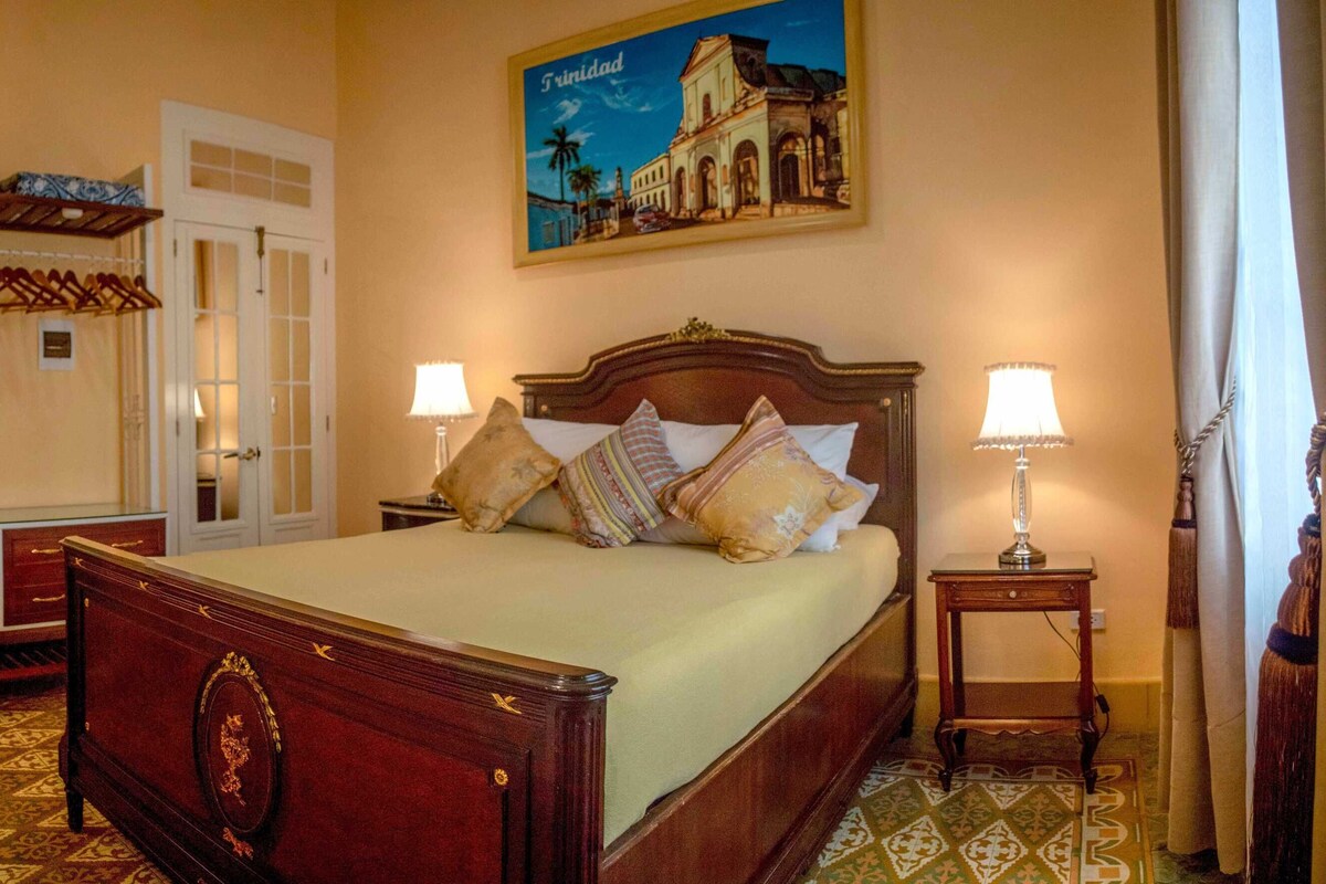 A hotel room with a large bed, two bedside tables, a wardrobe, a mirror, a television, and a painting of Trinidad.