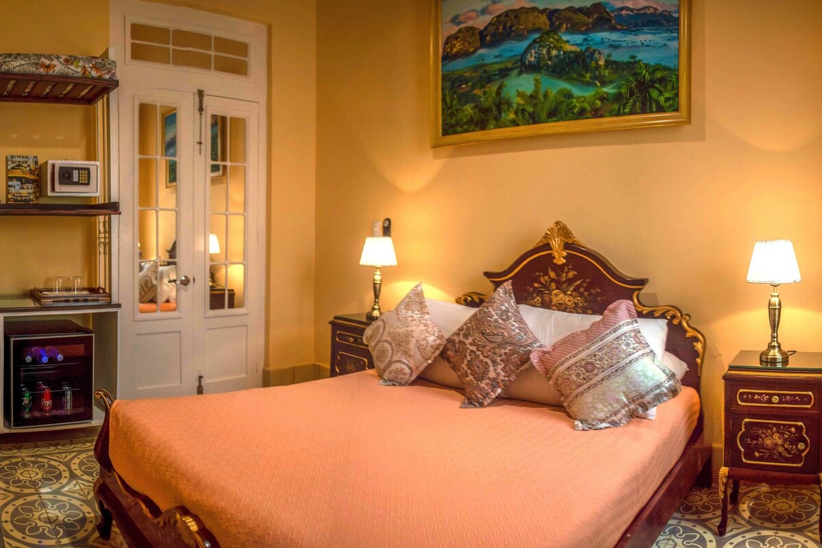 A hotel room with a bed, two bedside tables with lamps, a wardrobe, a microwave, a small cabinet, and a painting on the wall.