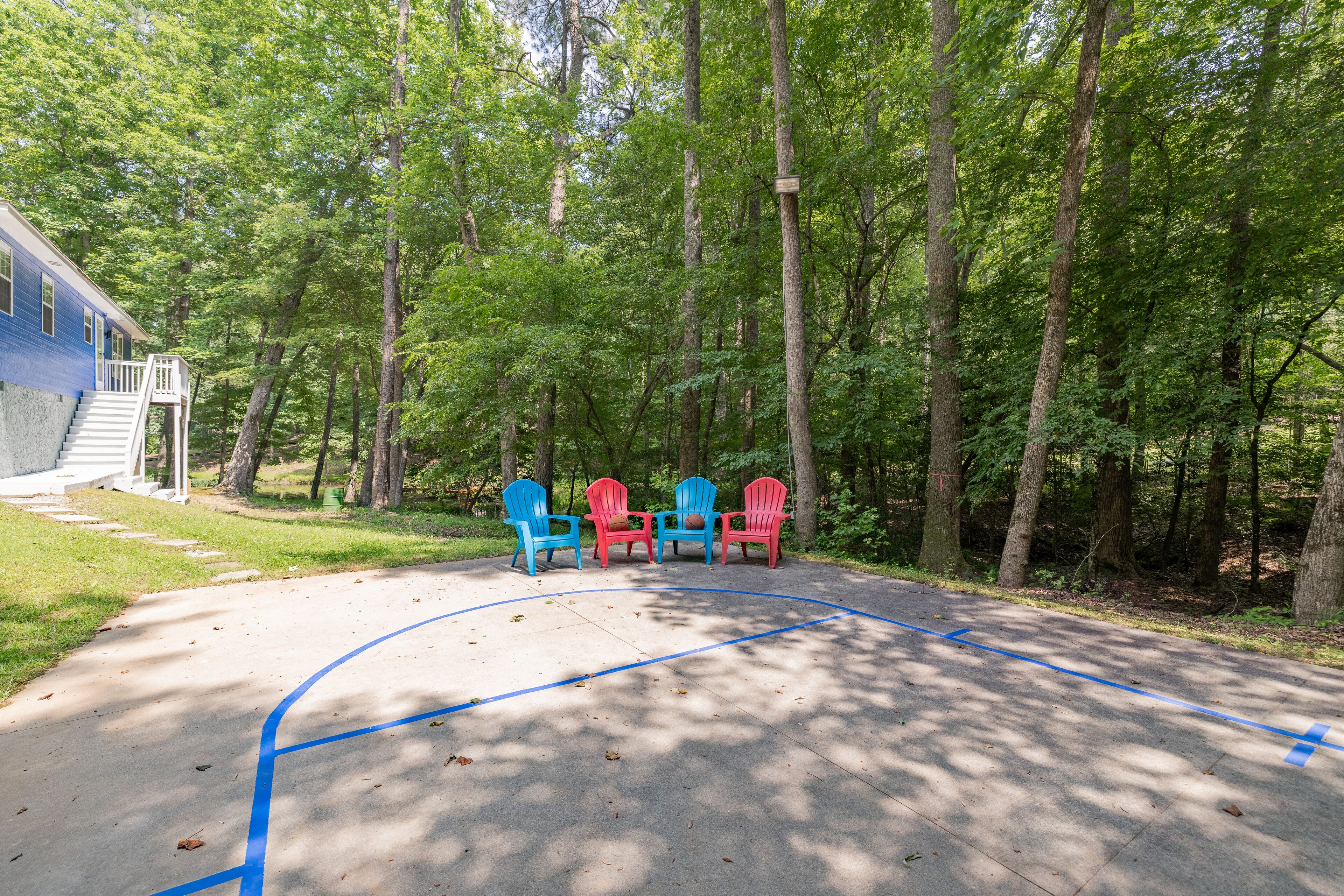 A basketball court with blue lines, surrounded by trees and three Adirondack chairs in blue, red, and gray.