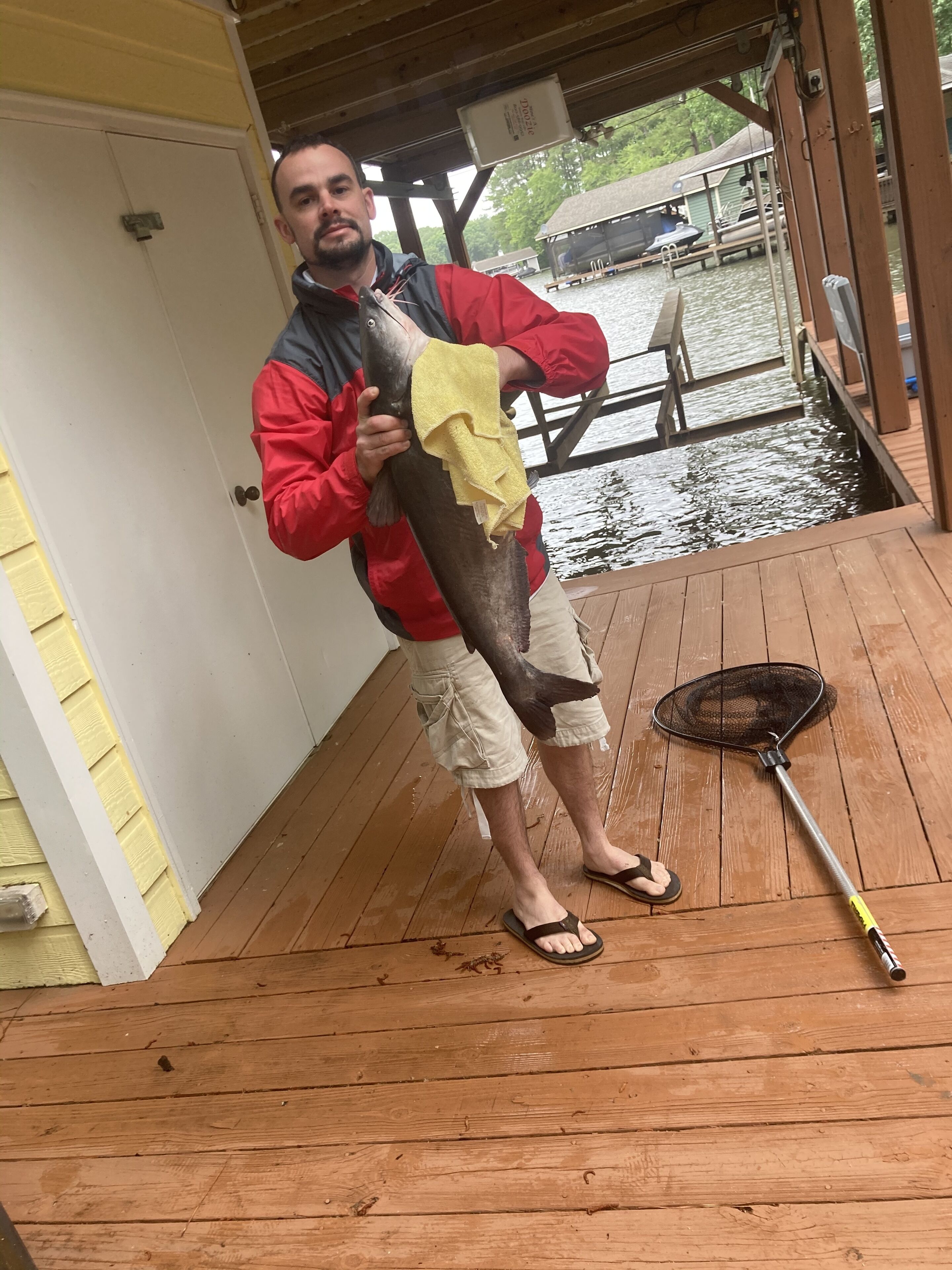 A person holding a large fish on a wooden dock with a fishing net and a boat in the background.