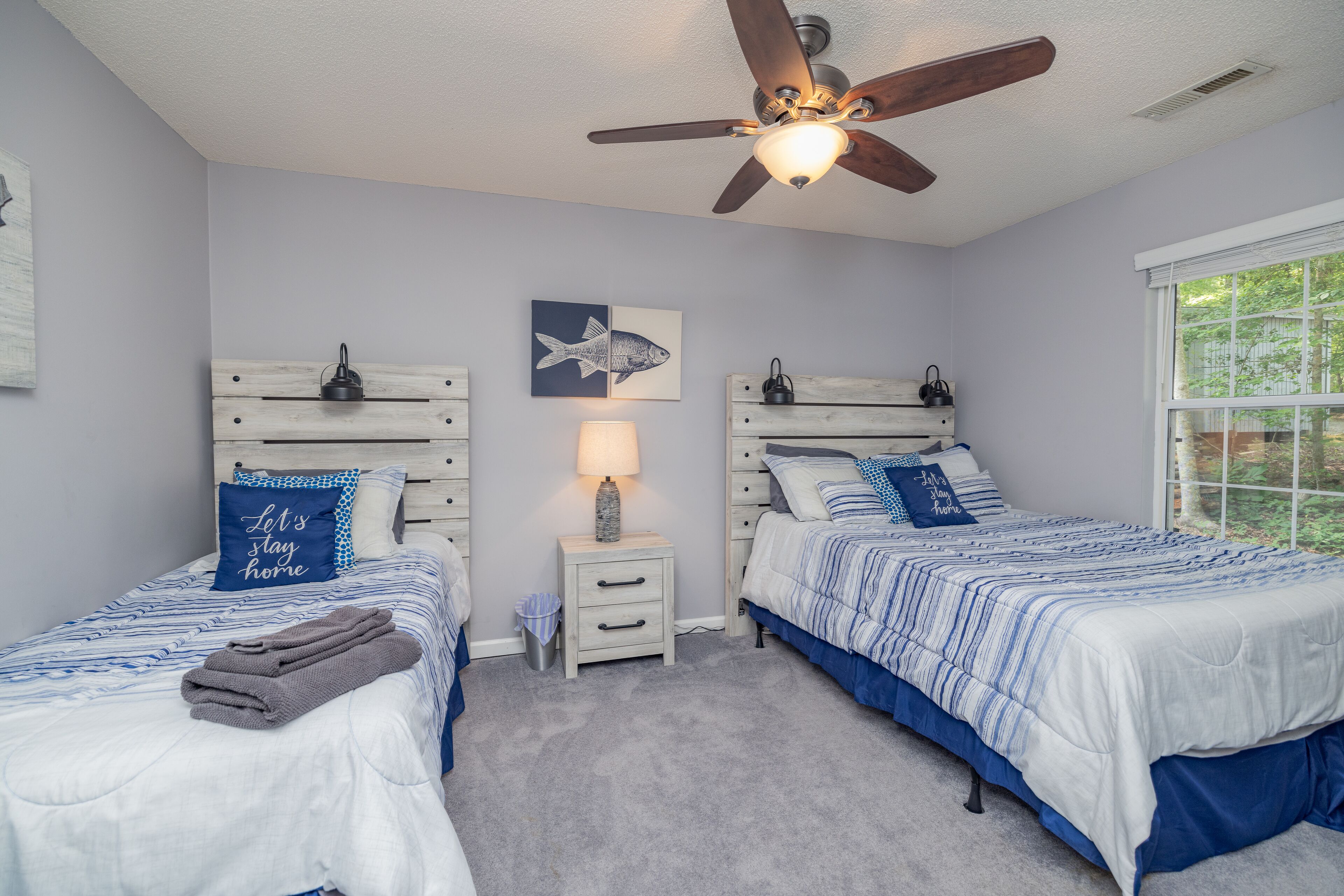 A bedroom with two beds, a ceiling fan, a window, and a nightstand with a lamp.