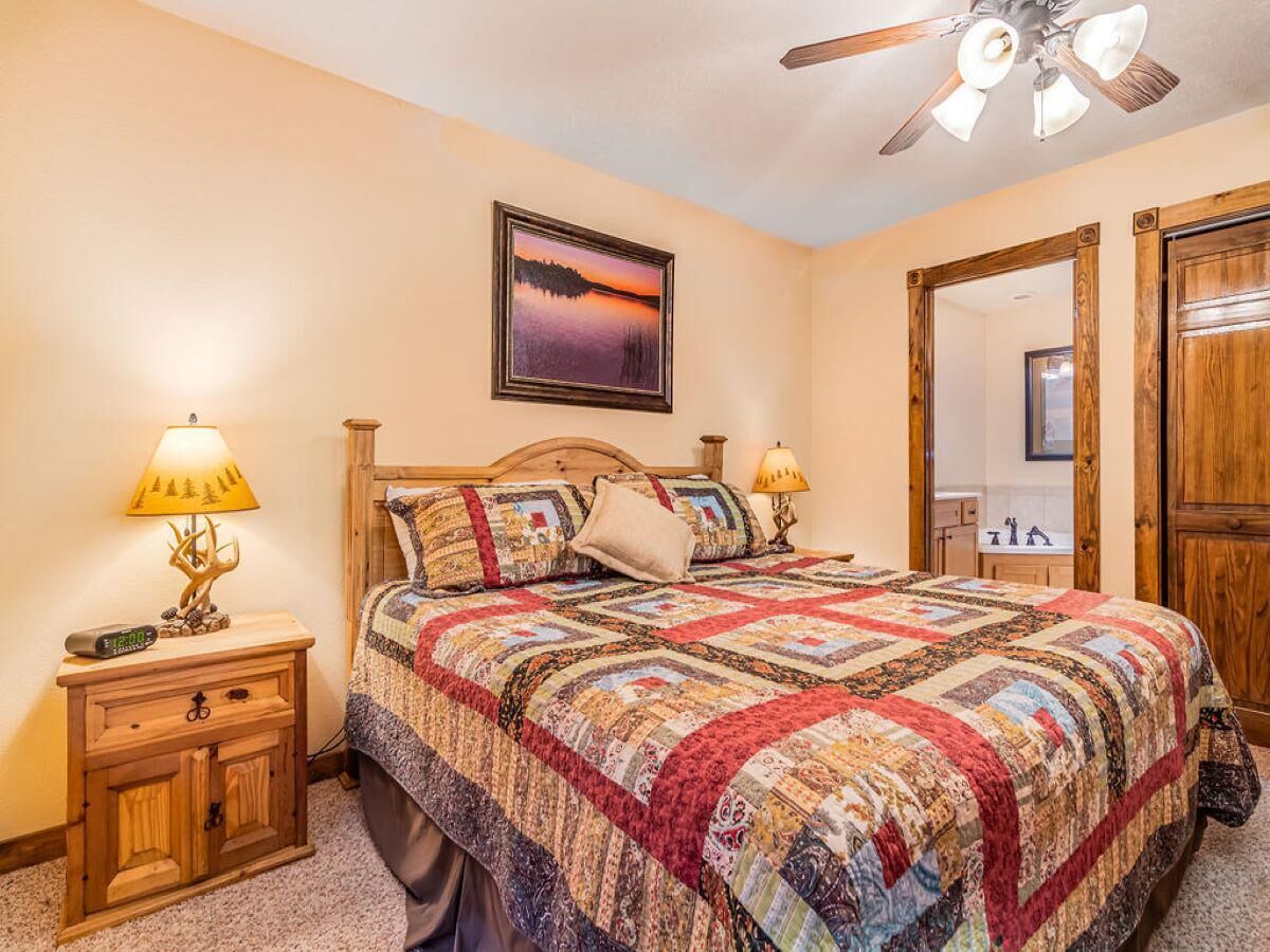 A bedroom with a wooden bed, a quilt, two bedside lamps, a nightstand, a ceiling fan, and a framed picture on the wall.