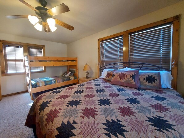 A bedroom with a bunk bed, a large bed with a patterned quilt, a ceiling fan, and wooden blinds. - Bear Hug (Reeds Spring)