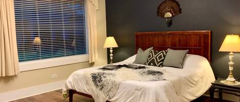 A bedroom with a large bed, two bedside tables with lamps, a window with blinds, a chandelier, and a decorative wall piece.