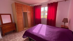 Ein Schlafzimmer mit einem Bett, einem Holzschrank, einem Spiegel, einem Fenster mit roten Vorhängen und einem Nachttisch mit einer Lampe.