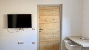 Ett rum med en dörr av trä, en väggmonterad tv, ett skrivbord med en stol och en vit vägg.