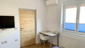 Ett sovrum med en säng, ett skrivbord, en stol, en tv som är monterad på väggen och ett fönster med persienner.