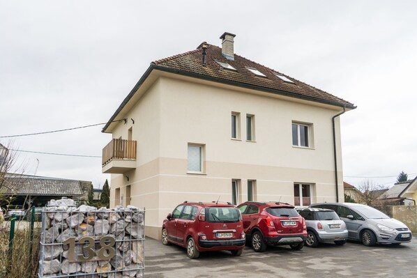 A two-story house with a tiled roof, a balcony, and a driveway with parked cars.