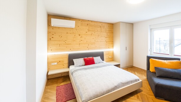 A modern bedroom with a wooden headboard, a bed with grey bedding, a red pillow, a grey sofa with a yellow pillow, and a window with a view of buildings.