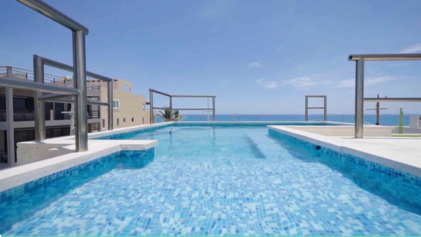 En swimmingpool med kristallklart blått vatten, omgiven av moderna byggnader och med utsikt över havet.