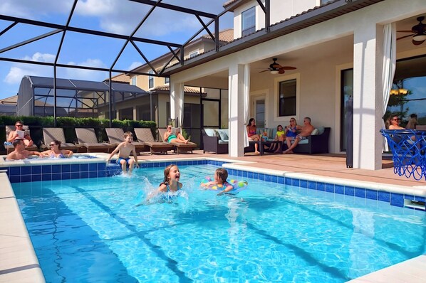 A swimming pool with people in it, a covered patio area, and a view of houses in the background.