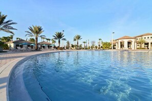 A swimming pool with lounge chairs and umbrellas, surrounded by palm trees and buildings.