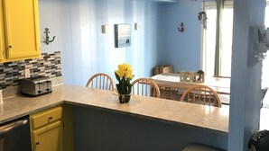 Une cuisine avec des meubles de couleur jaune, un grille-pain, un vase rempli de fleurs jaunes et un coin repas avec une table et des chaises.