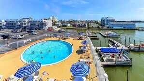Un espace piscine avec des chaises longues et des parasols, un quai avec des bateaux, et des bâtiments en arrière-plan.