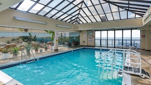 An indoor swimming pool with a glass ceiling, surrounded by potted plants and a view of the city. - Direct Oceanfront condo- AMAZING VIEWS & GREAT AMENITIES! (Ocean City)