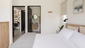 Una habitación de hotel moderna con una cama grande, una mesita de noche con lámpara, una ventana con vistas y un baño visible a través de una puerta abierta.