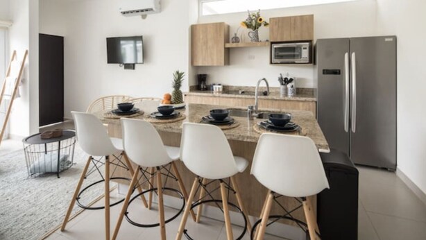 Una cocina moderna con una isla central, sillas blancas, un refrigerador de acero inoxidable y armarios de madera.