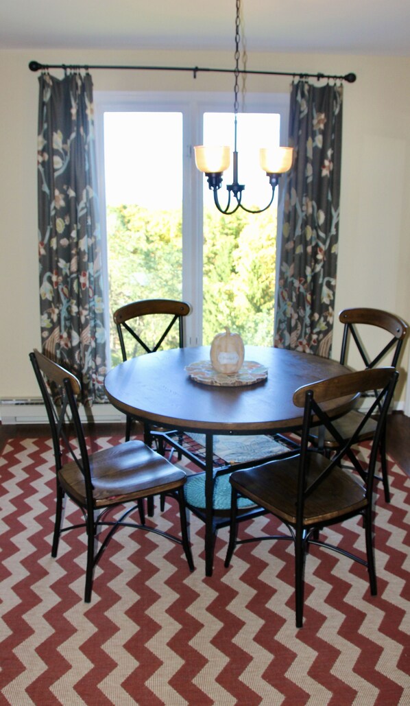A dining area with a round table, four chairs, a chandelier, and a view of greenery through the window.