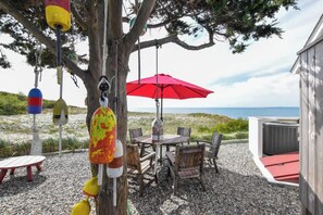 An outdoor seating area with a table and chairs, a red umbrella, and a view of the beach. - Beachfront on Private Beach w/ AC! (Truro)