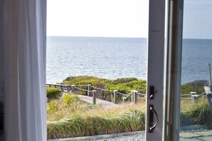 A view from a room with a glass door, overlooking a wooden deck leading to a body of water, with greenery and a clear sky.