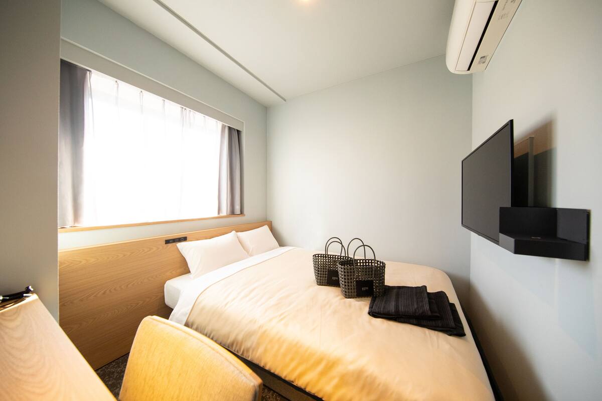 A hotel room with a bed, a TV mounted on the wall, a window with curtains, and a bedside table.