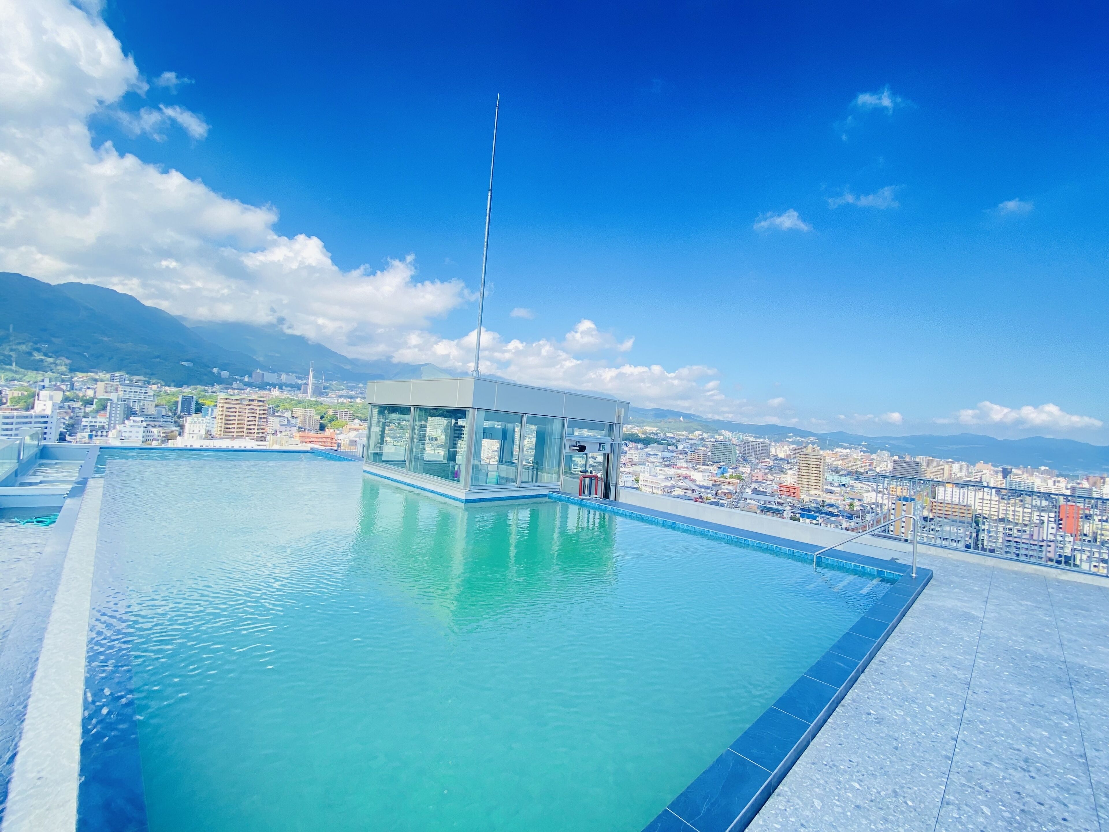 A rooftop infinity pool with a glass enclosure and a flagpole, overlooking a cityscape with mountains in the distance.