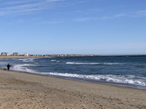 A beach with a person walking along the shore, waves crashing onto the sand, and a distant shoreline with buildings. - A Perfect Family Beach House! (South Kingstown)