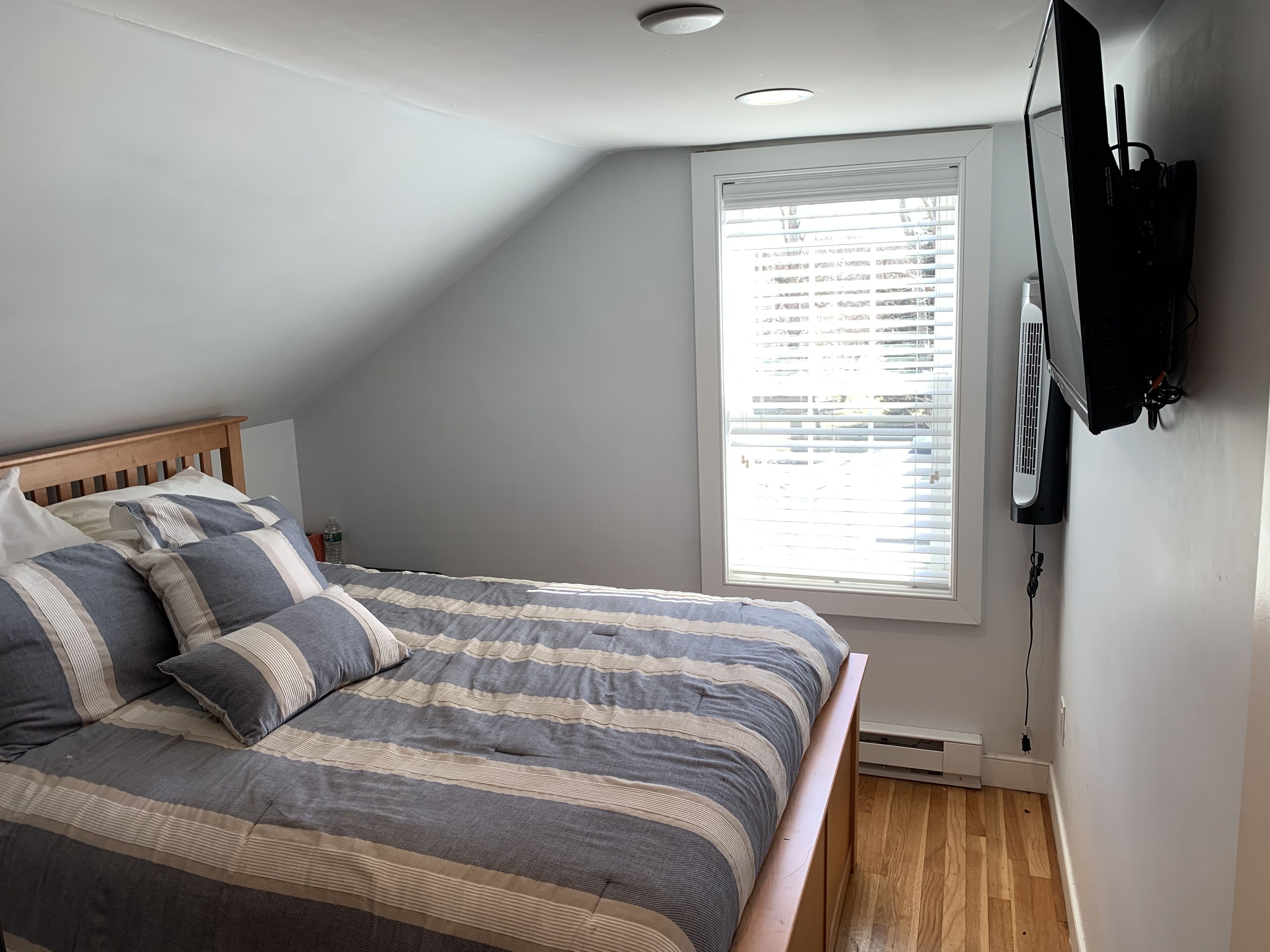 A bedroom with a bed, a window with blinds, a ceiling light, and a wall-mounted TV.