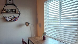 Una habitación pequeña y bien iluminada, con una mesa de madera, una silla morada, una lámpara de escritorio y una ventana con persianas.