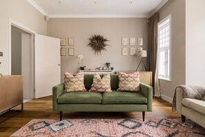 A living room with a green sofa, patterned cushions, a decorative wall piece, framed pictures, and a wooden floor.