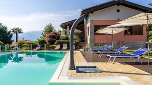 Piscina com espreguiçadeiras e guarda-sóis, rodeada por um edifício com vista para montanhas.