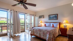 Ein Schlafzimmer mit einem Bett, zwei Nachttischlampen, einem Deckenventilator und Blick durch eine offene Tür auf einen Außenbereich mit Tisch und Stühlen.