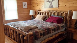 A wooden cabin bedroom with a bed, two bedside tables, a window, and a wall with a deer painting.