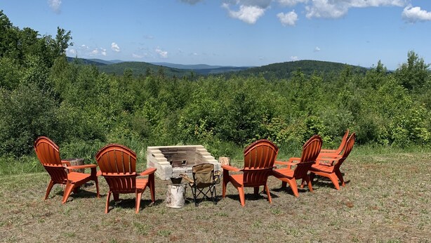An outdoor seating area with four orange chairs and a table, a fire pit, and a view of green hills under a clear sky.