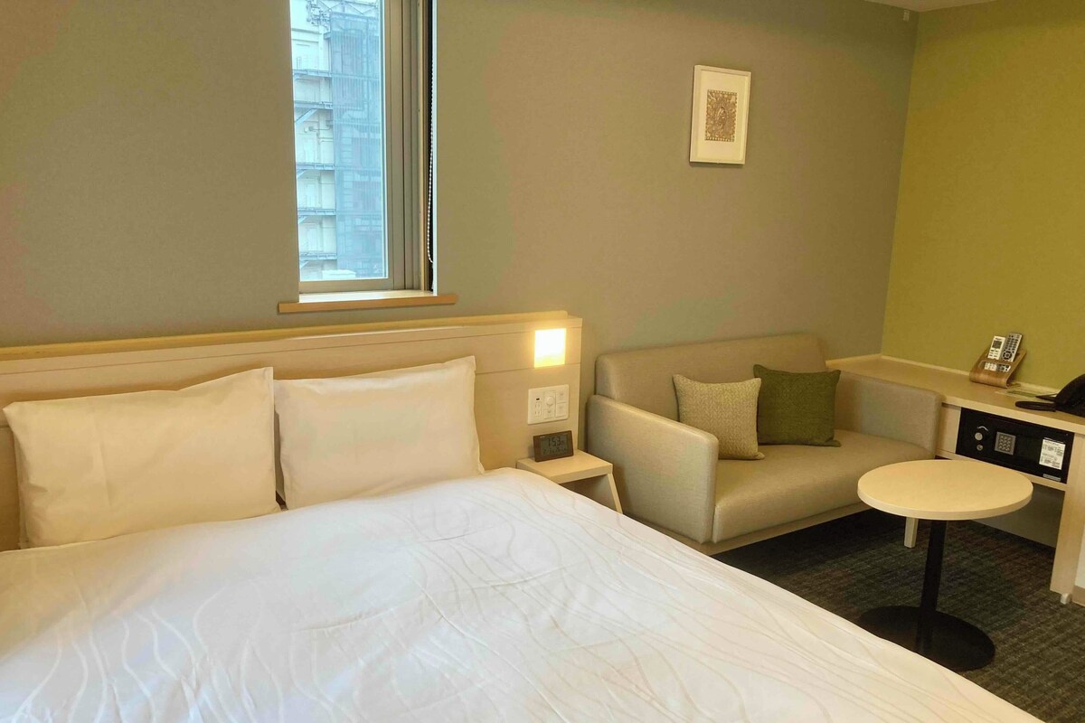 A hotel room with a large bed, a sofa, a small table, and a bedside table with a lamp and a phone.