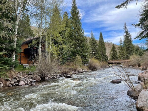 A cabin by a river with a wooden exterior, surrounded by trees and a rocky riverbank.