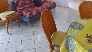 Un salon avec un canapé, deux fauteuils, une table à manger recouverte d’une nappe jaune, une cuisine visible à travers une arche, et une fenêtre avec des rideaux verts.