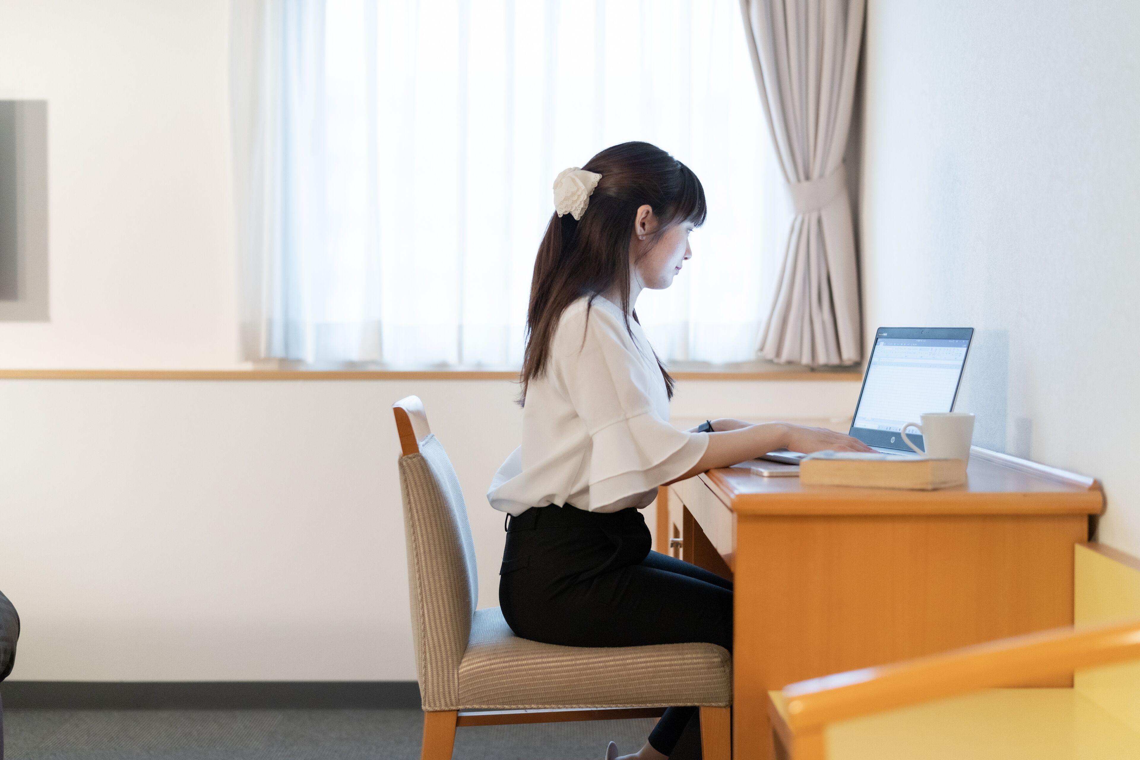Iemand zit aan een houten bureau met een laptop, een boek en een beker. De kamer heeft een raam met gordijnen, een stoel en een wandplank.