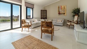 Un soggiorno spazioso con un'ampia porta-finestra scorrevole che si apre su una terrazza, un divano, una sedia in legno e una televisione.