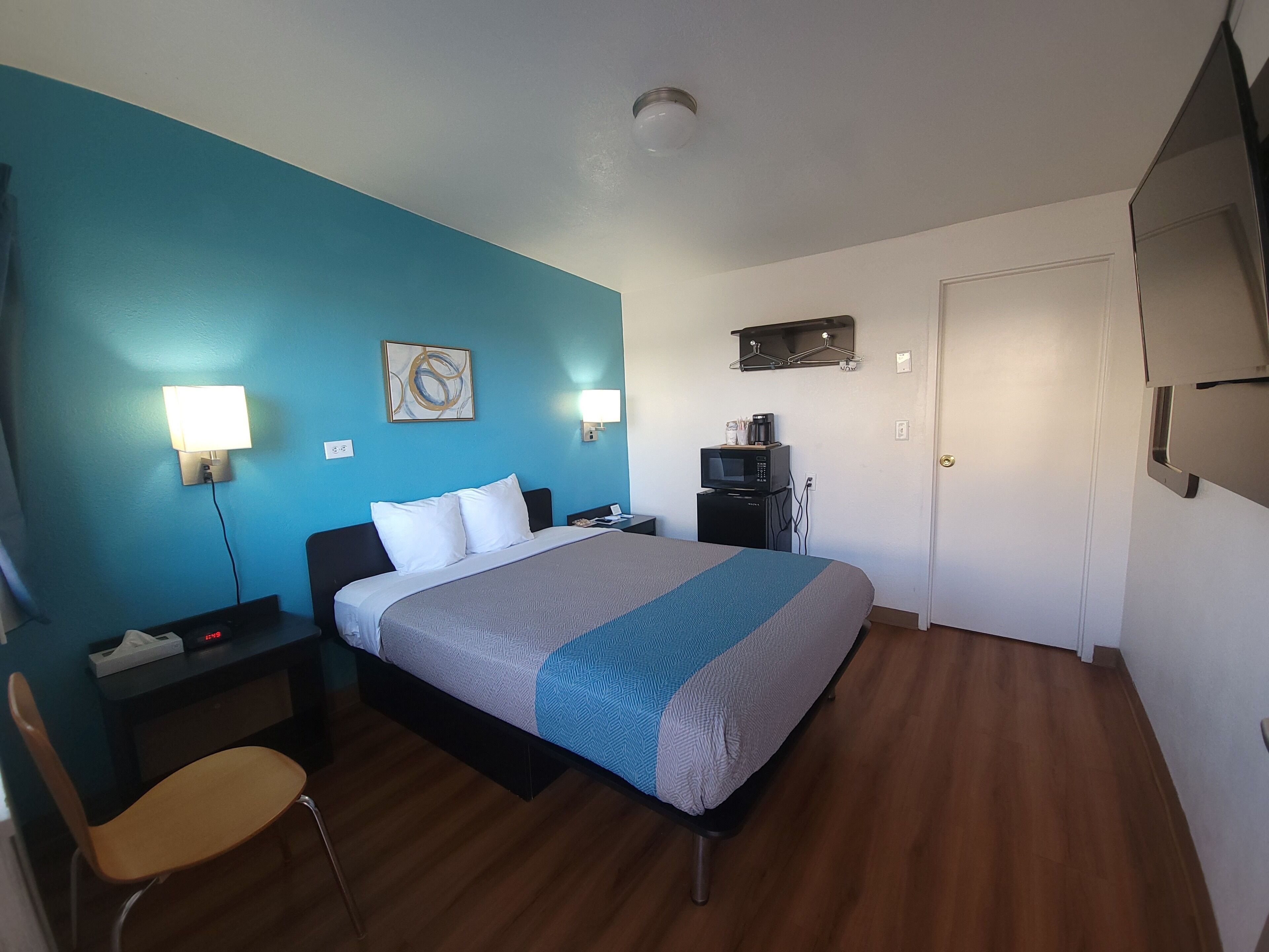 A hotel room with a bed, bedside table, chair, desk, and a microwave on a small cabinet.