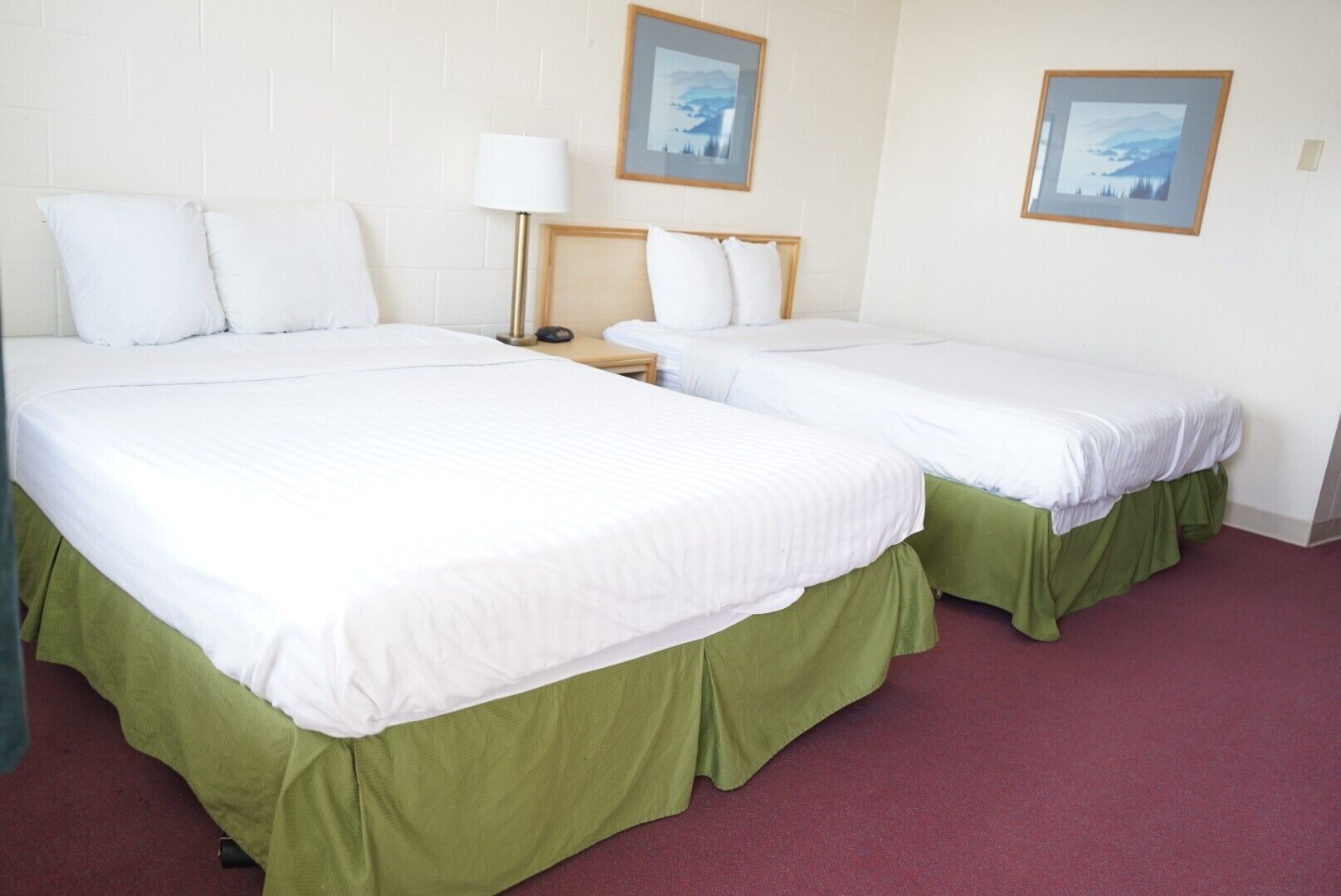Ein Hotelzimmer mit zwei Betten, jeweils mit weißen Laken und grünen Bettlaken. Zwei Nachttische, einer mit einer Lampe, und zwei gerahmte Bilder an der Wand.
