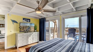A bedroom with a bed, a ceiling fan, a dresser, a painting, and a view of the outdoors. - Charming Beachfront Cottage Awaits You! Direct access to beach off deck/walkway. (Oak Island)