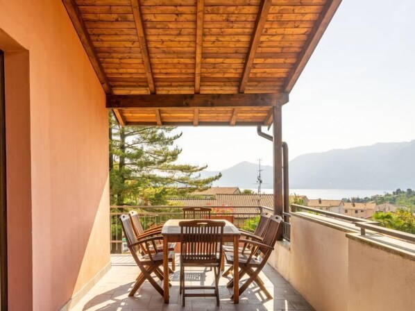 A wooden pergola-covered balcony with a table and chairs, overlooking a lake and mountains.