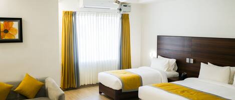 A hotel room with two beds, a sofa, a coffee table with fruit, and a ceiling fan.