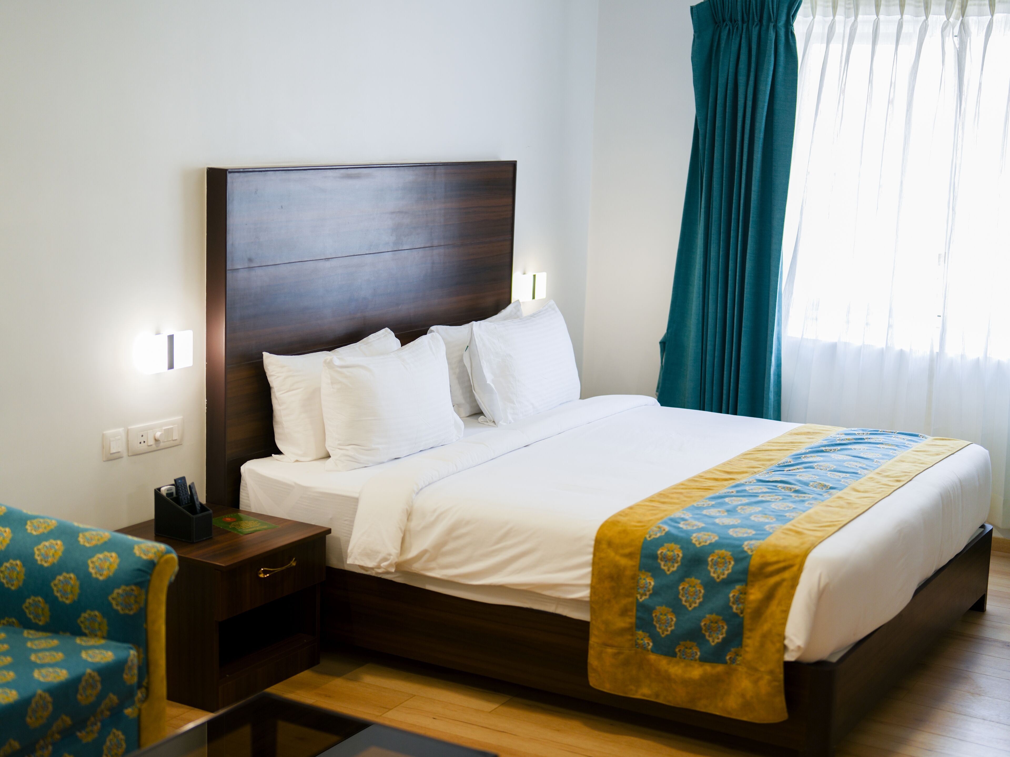 Una cama bien hecha con cabecera de madera, una mesita de noche con lámpara y una silla con cojín estampado.