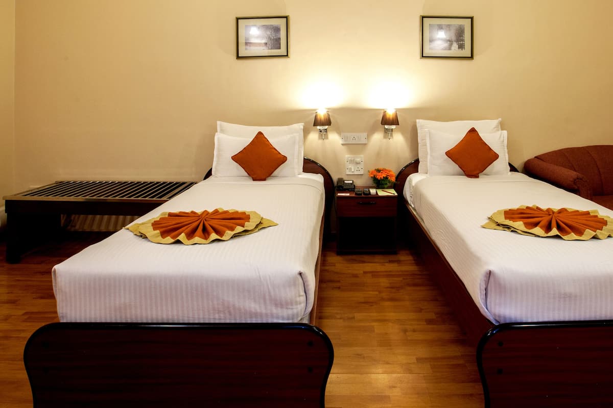Habitación de hotel con dos camas individuales, suelo de madera, dos lámparas de noche, una mesita de noche con un teléfono y un jarrón con flores, y un sillón rojo.