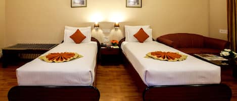A hotel room with two single beds, a wooden floor, two bedside lamps, a nightstand with a phone and a vase of flowers, and a red armchair.