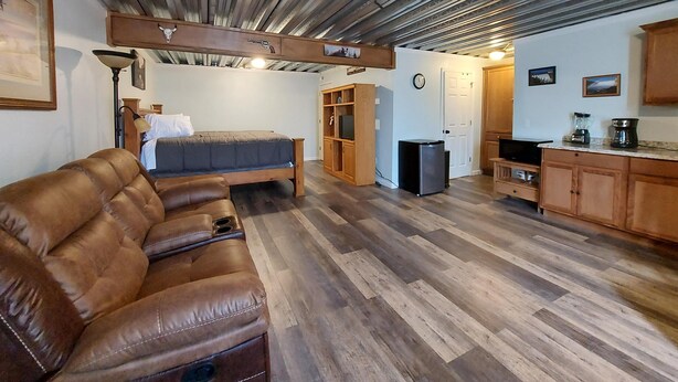 Een gezellige kamer met een bruine leren bank, een bed met een grijze deken, houten kastjes, een kitchenette met een koelkast, en een klok aan de muur.