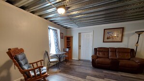 Een woonkamer met een houten schommelstoel, een bruine leren bank, een eettafel met stoelen, en een plafond met zichtbare balken.