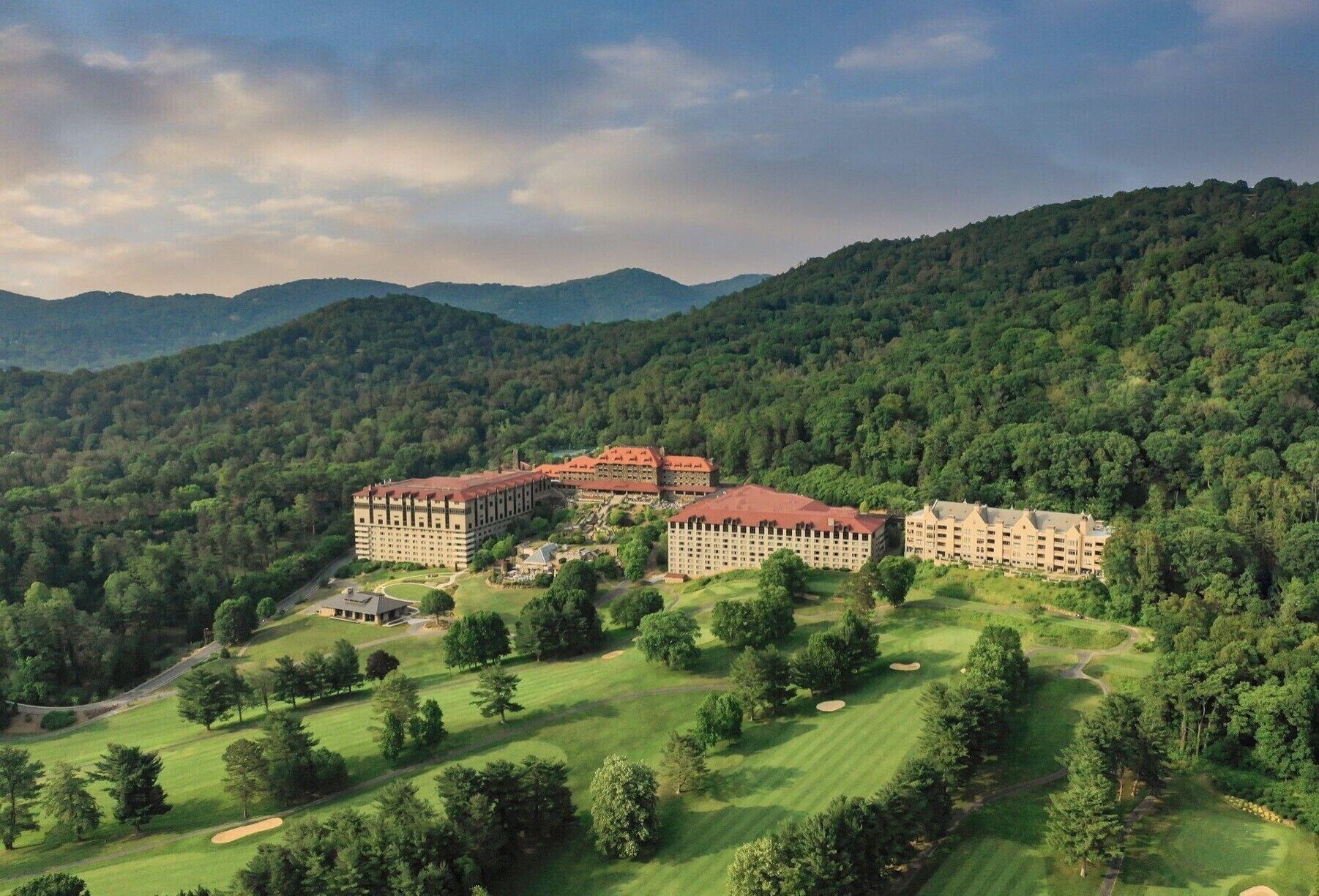 A large, multi-story hotel with a central courtyard, surrounded by lush green hills and a mountainous backdrop.